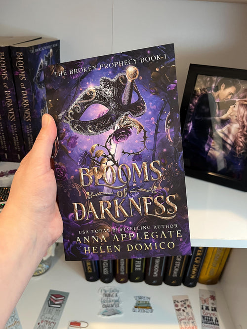 Book titled 'Blooms of Darkness' held by a hand with more books in the background.