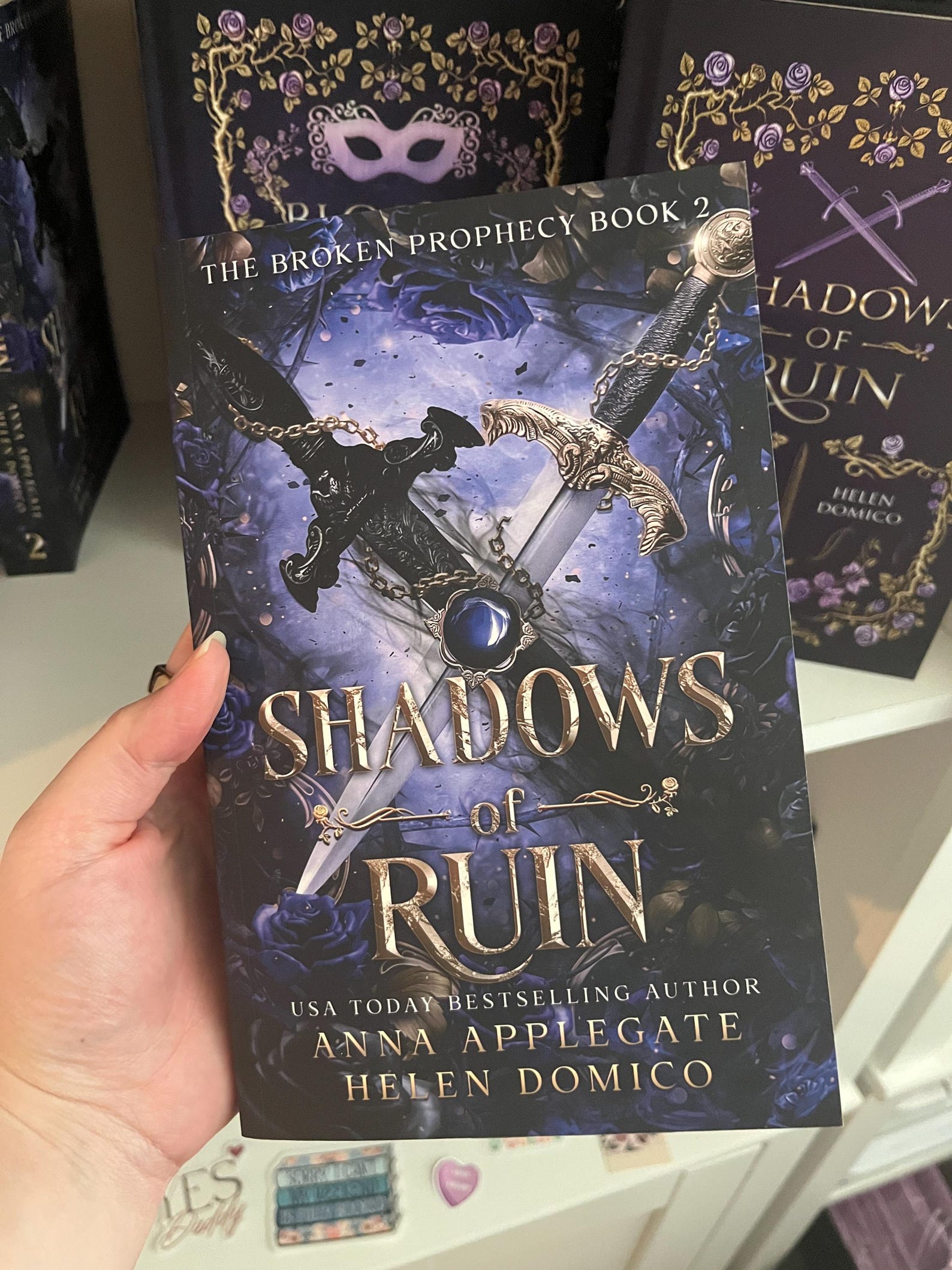 Book titled 'Shadows of Ruin' held by a hand with more books in the background.